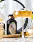 A clear, elegant coupe glass filled with a golden beverage sits in the foreground on a white table with an ornate white and gold glass to its left. The background features a blurred black circular frame, white flowers, and a decorative black apothecary jar hinting at the charm of Magic Hour's Pearl Tea of Beauty, Clarity, and a Sense of Peace.