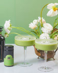 Two glasses of a green matcha drink are placed on a white surface. Beside them are two containers of organic matcha powder and syrup, with elegant black and green packaging. In the background, there is a decorative vase filled with white flowers against a green wall, embodying the essence of Magic Hour's Ceremonial Matcha Traveler Gift Box.