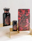 Three luxurious dark-hued containers with intricate red and gold geometric designs, labeled as Magic Hour's Gemstone Bundle: Garnet, Amethyst, Aquamarine, are elegantly displayed on golden stands against a light background. The products include a large bottle, a smaller jar, and a resealable bag.