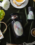 A luxurious flat lay featuring a tea set with a cup of Pearl Tea of Beauty, Clarity, and a Sense of Peace by Magic Hour, a black tea container, a small jar, a bowl of loose leaf tea with a spoon, white and green flowers, a string of pearls, a folded fan, and decorative items on a dark green velvet background.