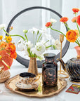 A decorative setup featuring a Magic Hour Scorpio Tea for Sensual Brilliance tin, vintage teacup and saucer, black teapot, vases with white and orange flowers, and a gold-trimmed tray. Additional items include a gold watering can, a small round mirror, and a wine glass, all set on a white surface.