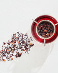 A glass cup filled with vibrant Magic Hour Immune Boosting Teas Sampler Set sits on a white surface. Loose leaf tea and petals are scattered beside the cup, while a metal tea infuser containing organic tea is placed on top, casting a delicate shadow.
