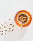 A glass cup of tea with a tea infuser placed inside, containing organic tea leaves and spices from the Rest, Relaxation & Sleep Sampler Set by Magic Hour. Loose spices, including cloves and cardamom pods, are scattered on a white surface next to the cup. The image has a bright and clean aesthetic.