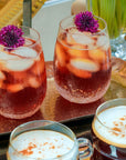 Two glasses of dark iced beverages garnished with purple flowers sit on a copper tray. Nearby are two small glass cups filled with frothy, creamy drinks topped with cinnamon. The mood-boosting teas from the Coffee Lovers Tea Kit for Better Moods, Digestion & Metabolism create a fresh, inviting ambiance, complemented by green plants and drinkware in the blurred background.