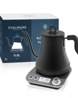 A black Magic Hour Electric Pour Over Kettle with a gooseneck spout is displayed on a built-in base with a digital screen and control dial, perfect for brewing loose leaf tea. The kettle packaging, showing a similar image and branding, is positioned behind the kettle. The text on the packaging reads "Magic Hour.