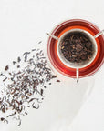 A glass cup filled with brewed organic tea sits on a white surface. A metal tea infuser holding loose leaf Teas for Toning, Slimming & Weight Release Sampler Set by Magic Hour rests on top of the cup. Some loose tea leaves are scattered on the surface next to the cup, casting a shadow.
