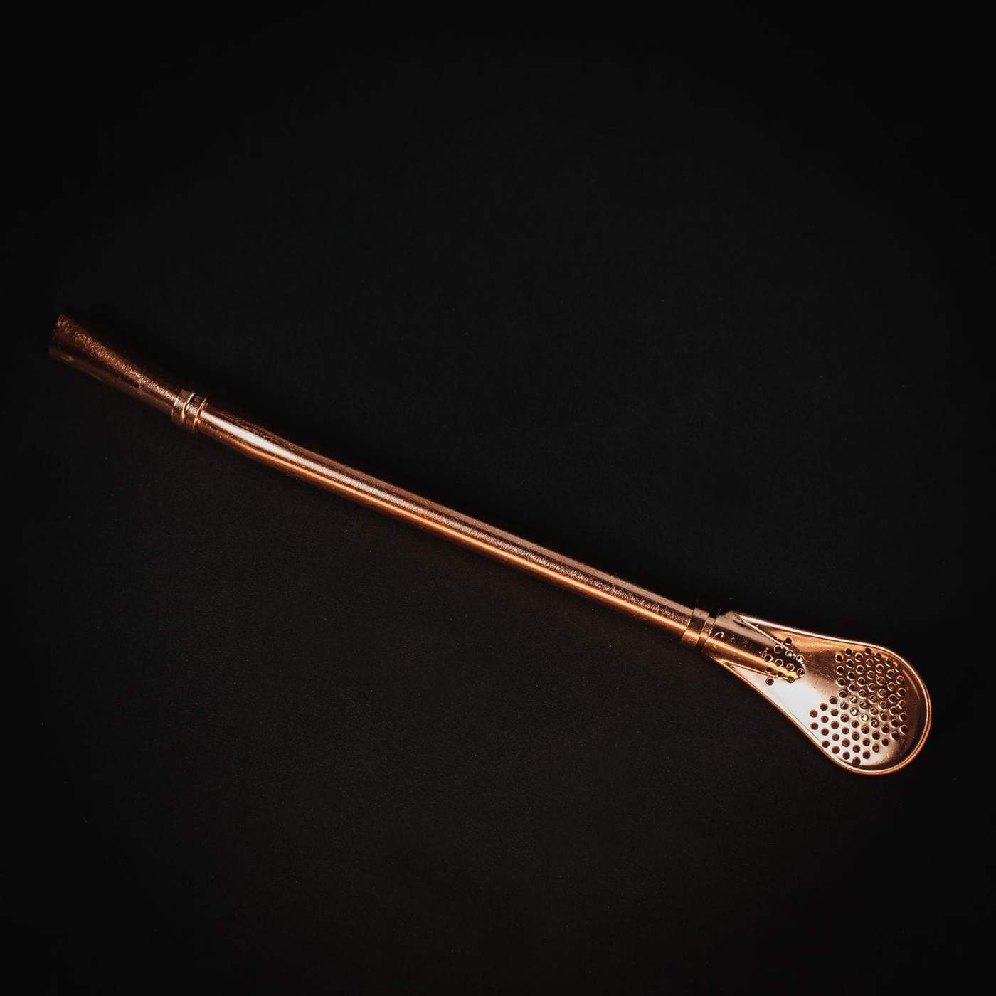 A Magic Hour Sip Wellness: Ritual Kit for Immunity & Joy, which includes a brass or copper bombilla—a type of straw used for drinking mate or organic tea—is placed on a dark background. The bombilla features a cylindrical stem that widens at the end with a perforated, spoon-like filter. The metal surface has a polished, reflective finish.