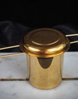 A gold-colored Luxury Traveler Gift Box from Magic Hour, designed for loose leaf tea, boasting a cylindrical mesh body and two extended handles, rests elegantly on a white marble slab. The strainer's lid matches in gold color, and the black background adds a touch of sophistication.