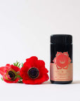 A container of Magic Hour Blissful Black Teas Medley with a pink label reading "Sacral Chakra Steam Blend - Joyous" is placed on a white surface. Two bright red flowers with dark centers lie next to the container. The background is plain white.