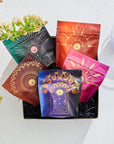 A collection of colorful, decorative tea packages displayed in a black box. Featuring intricate, gold-embossed designs in green, black, orange, red, and purple colors. The Luxe Tea Sampler Sips: The Queens Collection by Magic Hour is set on a white surface with flowers and a glass of water in the background.