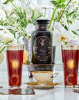 A decorative scene with a large black apothecary jar labeled "Virgo Tea of Virtue, Wit & Meticulous Magic" surrounded by white flowers. Two tall pinkish glasses with sun designs and a teacup of Magic Hour tea are placed on the table. Elegant candle holders and floral arrangements complete the setting.