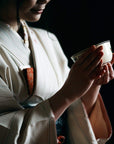 A person dressed in a traditional Japanese kimono is holding a ceramic bowl of Kansai-Kyoto Ceremonial Matcha by Magic Hour with both hands. The image is dimly lit, focusing on the serene and delicate gesture, with the individual’s face partially visible.