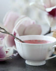 A white teacup filled with pink liquid, placed next to a silver spoon. Pink and white magnolia flowers and a book are also visible in the background, creating a serene and delicate ambiance—a perfect setting for enjoying Magic Hour Magnolia Rose Oolong Tea.