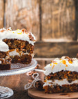 A single slice of Year of the Rabbit Carrot Cake with cream cheese frosting and chopped nuts sits on a wooden board, accompanied by a steaming cup of herbal tea. The rest of the cake is on a glass cake stand, against a rustic wooden background.