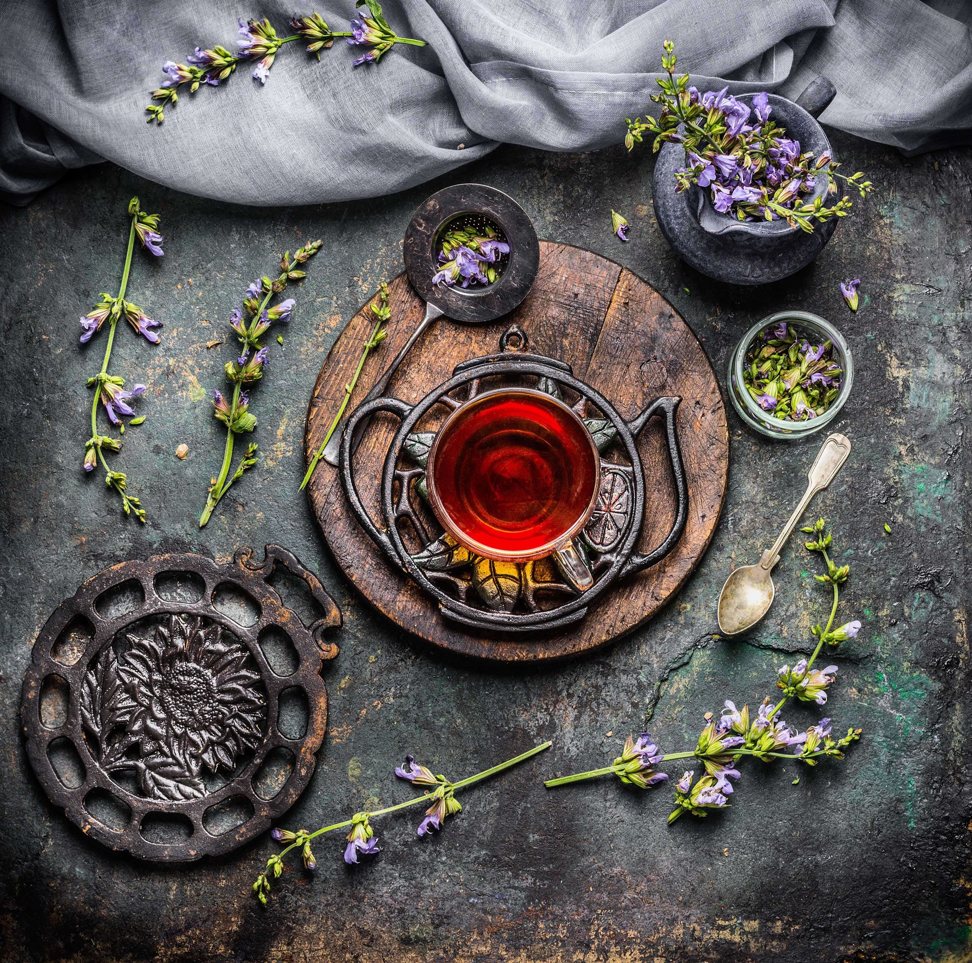 A rustic tea setup features a cup of Self-Guided 14 Day Ceremonial Sensory Tea Cleanse Kits by Club Magic Hour on a wooden board surrounded by a black metal trivet, a spoon, and various flowers. A grey cloth is draped above, with small purple flowers scattered around, lending a natural and vintage aesthetic.