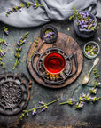 A rustic tea setup features a cup of Self-Guided 14 Day Ceremonial Sensory Tea Cleanse Kits by Club Magic Hour on a wooden board surrounded by a black metal trivet, a spoon, and various flowers. A grey cloth is draped above, with small purple flowers scattered around, lending a natural and vintage aesthetic.