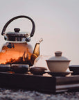 A glass teapot filled with Sin Eraser™ : Puerh Tea from Magic Hour sits on a wooden tray alongside several small cups and bowls. The tea has a warm amber hue, and the setup is outdoors, suggesting a serene, peaceful atmosphere.