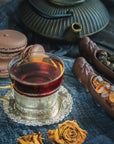 A glass cup of Organic Golden Monkey Tip Yunnan Black Tea by Magic Hour sits on a silver saucer, surrounded by brown macarons, a dark teapot, dried roses, and dishes holding rock sugar and loose leaf tea. All items are arranged on a textured blue cloth.