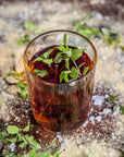 A glass of dark brown iced Bliss: Sacred Tulsi Adaptogen Tea from Magic Hour garnished with fresh mint leaves sits on a surface sprinkled with coarse salt and scattered green herbs. The translucent drink reveals the mint leaves, giving it a refreshing and earthy appearance.