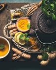 A rustic flat lay of a tea setting featuring Club Magic Hour's Self-Guided 14 Day Ceremonial Sensory Tea Cleanse Kits, a cup of organic tea garnished with mint, fresh turmeric root, sliced lemon, ginger, a bowl of turmeric powder, a honey dipper, and assorted herbs and loose leaf tea on a woven mat with wooden accents.