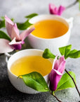 Two cups of Magic Hour Magnolia Oolong Tea with a light golden hue sit on a textured gray surface, surrounded by green leaves and vibrant purple-pink flowers, creating a serene and inviting setting.