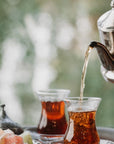 A close-up of two traditional glass cups filled with Magic Hour's Tea of The Rising Sun : Japanese Breakfast on a tray. A silver teapot is pouring tea into one of the cups. In the foreground, there are pieces of colorful Turkish delight. The background is blurred, showing green foliage.