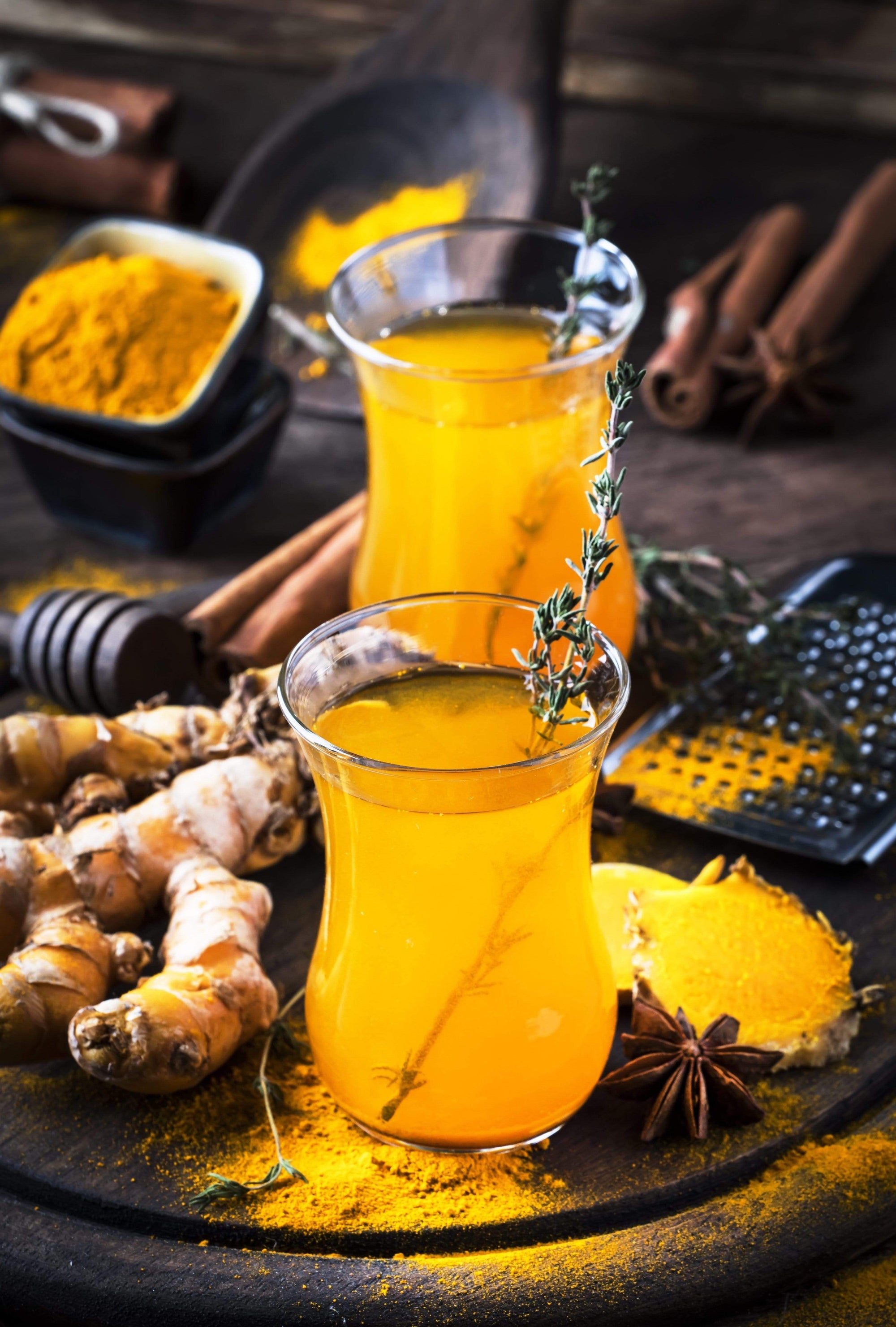 Two glasses of golden turmeric ginger tea with a sprig of thyme as garnish sit on a rustic wooden surface. Surrounding the glasses are fresh turmeric roots, a bowl of turmeric powder, cinnamon sticks, a honey dipper, and star anise—creating a cozy setting perfect for enjoying Club Magic Hour's Self-Guided 14 Day Ceremonial Sensory Tea Cleanse Kits.