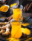 Two glasses of golden turmeric ginger tea with a sprig of thyme as garnish sit on a rustic wooden surface. Surrounding the glasses are fresh turmeric roots, a bowl of turmeric powder, cinnamon sticks, a honey dipper, and star anise—creating a cozy setting perfect for enjoying Club Magic Hour's Self-Guided 14 Day Ceremonial Sensory Tea Cleanse Kits.