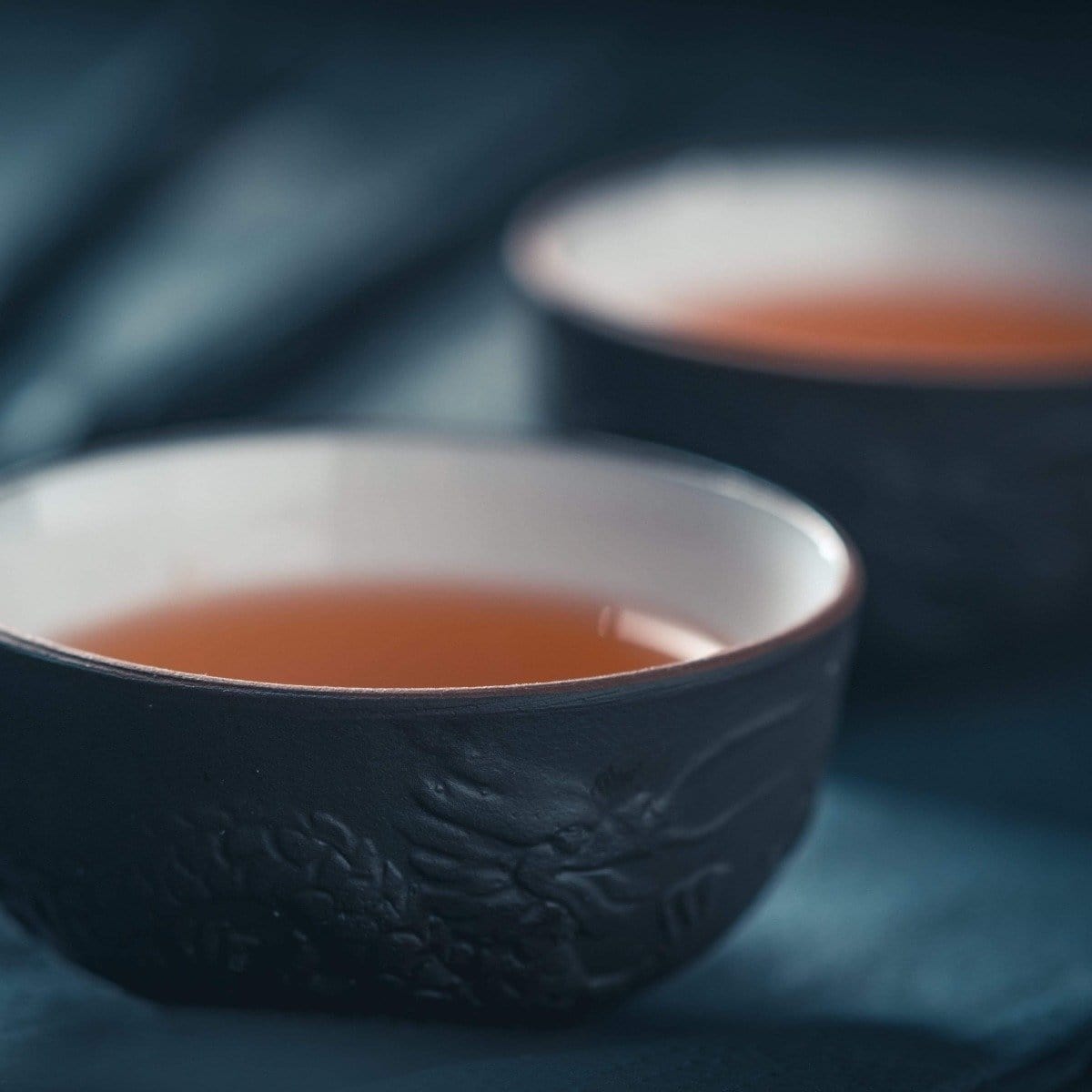Close-up of two ceramic cups filled with Renewal: Peach-Goji-Rose Oolong Tea Traveler by Magic Hour on a dark, blurred background. The cups have intricate designs, and the focus is on the front cup, emphasizing its texture and the warm color of the loose leaf tea.