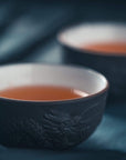 Close-up of two ceramic cups filled with Renewal: Peach-Goji-Rose Oolong Tea Traveler by Magic Hour on a dark, blurred background. The cups have intricate designs, and the focus is on the front cup, emphasizing its texture and the warm color of the loose leaf tea.