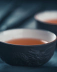 Close-up of a dark ceramic bowl filled with a warm brown liquid, likely Ti Quan Yin Oolong: Tea of Patient Compassion from Magic Hour, set against a blurred dark background. Another similar bowl is partially visible in the distance, suggesting a serene, contemplative setting that highlights the wellness benefits of this ancient brew.