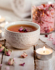 A delicate white ceramic cup filled with herbal tea, garnished with dried rosebuds, sits on a whitewashed wooden surface. Surrounding the cup are lit tea candles, scattered dried rose petals, and a glass jar containing Pink Tourmaline Bath Soak.