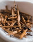 A white bowl filled with thin, dried brown tea leaves, arranged in a casual, slightly intertwined manner. This Organic Golden Monkey Tip Yunnan Black Tea from Magic Hour promises a rich infusion. The blurred background highlights the texture and details of these organic tea leaves.