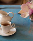 A white teacup filled with Magic Hour Magnolia Oolong Tea sits on a matching saucer alongside a white creamer on a blue table. Next to the tea setup, there is a small vase with pink and white flowers. The background is blurred, focusing on the items on the table.
