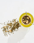 A glass teapot filled with yellow organic tea has a strainer on top containing a mix of loose leaf tea leaves and slices of almonds. Some tea leaves have spilled onto the white surface next to the teapot. The image is taken from above, showcasing the contents clearly. **Classics - Luxe Sampler Sips by Magic Hour** is beautifully presented in this image.