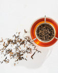Aerial view of a glass cup filled with amber-colored tea, showcasing The Astrology Collection - Luxe Sampler Sips. Loose rare teas are both in a tea infuser resting in the cup and scattered on the white surface beside it. The shadow of the infuser adds depth to the composition.