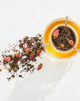 A clear glass teacup filled with amber-colored tea sits on a white surface. Beside it, there is a pile of The Astrology Collection - Luxe Sampler Sips' rare teas, adorned with pink rosebuds scattered across the surface. The teacup, designed by a master blender to enhance optimal flavor, features a sleek metal handle. The scene is brightly lit and minimalist.