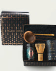 A gift box titled "Matcha 1111 : Gift Set" by Magic Hour is open to reveal its contents: a metallic tea strainer, a black tin with a colorful label, organic loose leaf tea, a straw whisk, and a black cylindrical container. The box and lid are black with a pattern of gold stars.