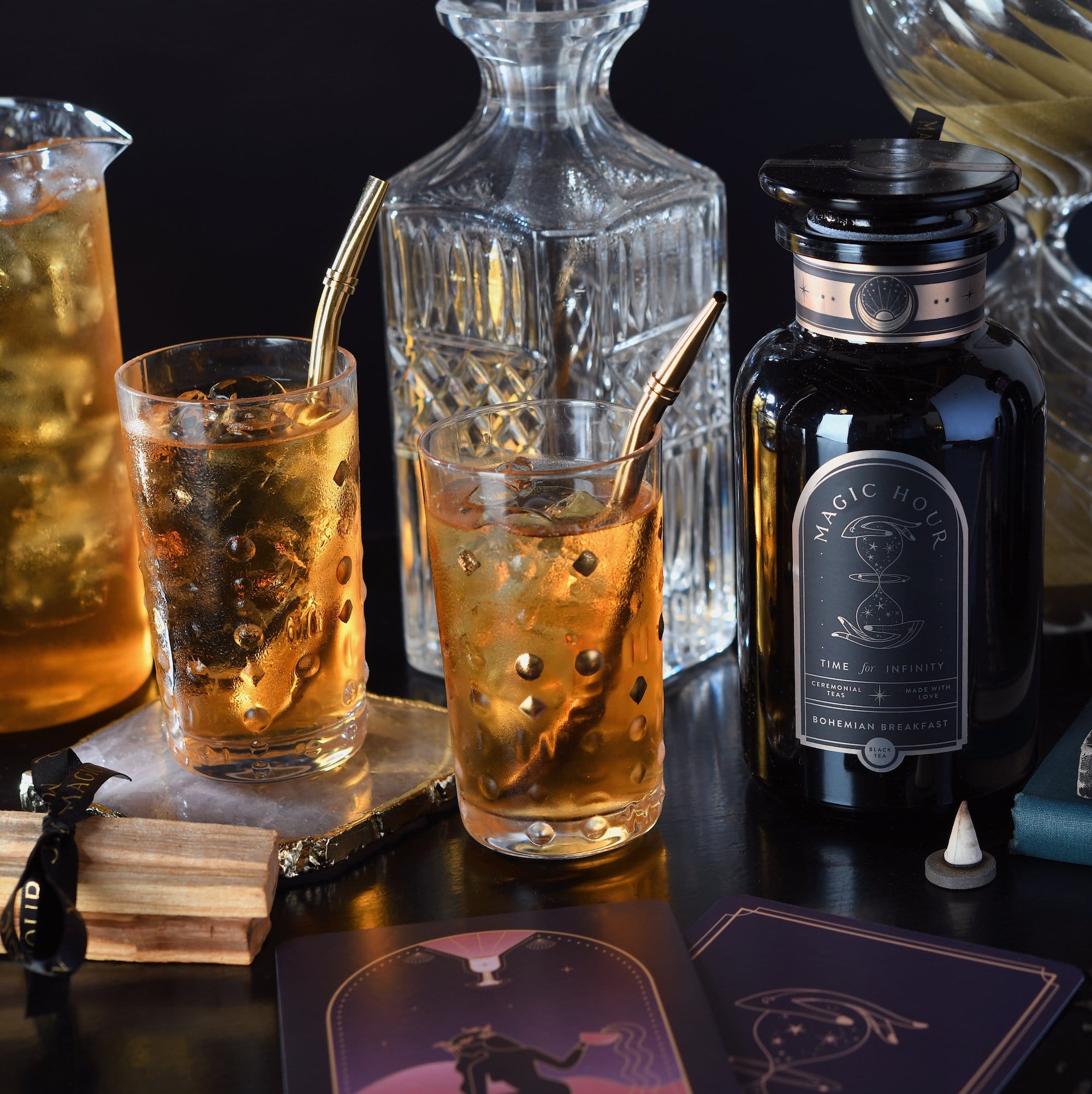 A crystal decanter, a black bottle of the Best Sellers of Magic Hour Box labelled "Magic Hour," and two glasses filled with an iced tea drink are arranged on a table. Gold boba straws are in the glasses, with tarot cards and a small incense holder nearby, creating an enchanting atmosphere.