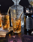 A crystal decanter, a black bottle of the Best Sellers of Magic Hour Box labelled "Magic Hour," and two glasses filled with an iced tea drink are arranged on a table. Gold boba straws are in the glasses, with tarot cards and a small incense holder nearby, creating an enchanting atmosphere.
