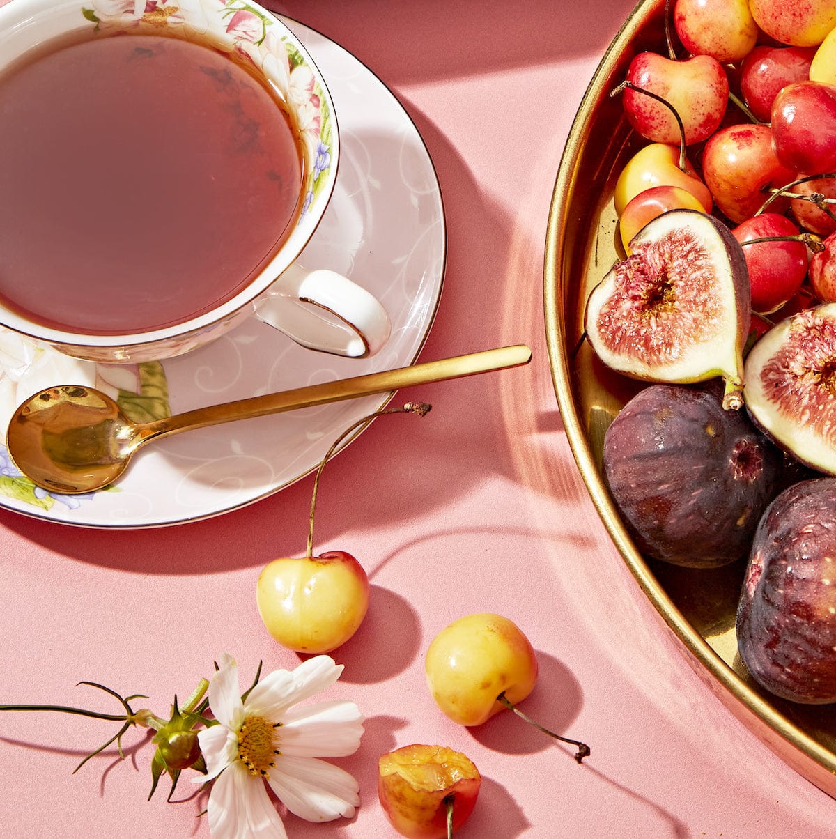 A delicate porcelain teacup from the Flower & Fruit Lovers Starter Set, filled with adaptogenic tea and accompanied by a gold spoon, sits on a pink surface. Nearby, a golden tray holds an assortment of fresh fruits, including figs and cherries. The scene is beautifully accented with a white flower and additional cherries scattered around.