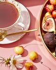 A delicate porcelain teacup from the Flower & Fruit Lovers Starter Set, filled with adaptogenic tea and accompanied by a gold spoon, sits on a pink surface. Nearby, a golden tray holds an assortment of fresh fruits, including figs and cherries. The scene is beautifully accented with a white flower and additional cherries scattered around.