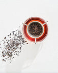 A glass teacup filled with dark, organic tea sits on a white surface. A tea infuser containing loose leaf tea rests on the cup, with additional loose tea leaves scattered to the left of the cup. The scene is brightly lit, casting soft shadows. The tea being enjoyed is part of Magic Hour's Classics - Luxe Sampler Sips collection.