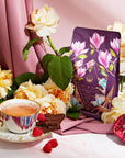 An elegant setup featuring a cup of tea on a saucer, a few pieces of chocolate, raspberries, half of a pomegranate, and a purple bag of **Breakfast in Paris-Oolong Black Tea Scented with Fruits & Flowers of Secret Parisian Gardens** adorned with floral designs. The scene evokes the charm of mystic Paris, decorated with yellow and pink flowers and luxurious fabric in the background.