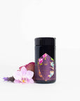 A dark-colored cylindrical container of "Breakfast in Paris-Oolong Black Tea Scented with Fruits & Flowers of Secret Parisian Gardens" sits against a white background. The container features a colorful geometric label with botanical designs reminiscent of Parisian gardens. Beside it is an arrangement of flowers, including a pink orchid and a red rose, adding a touch of elegance.