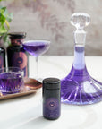 A Solstice Moon™-themed vignette featuring a glass carafe filled with purple liquid, a purple cocktail in a coupe glass, an organic tea blend in a tumbler on a copper tray, and a small black container with purple labeling, all arranged on a white countertop.