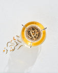 A glass tea infuser filled with the Astrology Bundle: Cancer, Leo, Virgo tea blend sits in a cup of brewed tea. Some loose tea leaves are scattered to the left on a white textured surface. The image is lit from the side, casting a shadow.