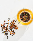 A top-down view of The Astrology Collection - Luxe Sampler Sips glass teapot filled with brewed rare teas. A metal infuser with dried tea leaves sits inside the teapot. Loose tea leaves are scattered beside the teapot on a white surface. Sunlight casts a soft shadow, adding a warm, cozy ambiance.