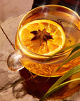 A clear glass teacup from the Spice Lovers Starter Set is filled with a golden liquid, garnished with a slice of orange and a star anise, resting on a glossy tiled surface. A green leaf partially frames the right side of the image, adding a touch of nature to the warm, cozy scene.