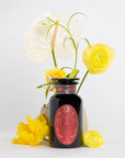 A decorative jar with a red label sits in front of a floral arrangement featuring white anthurium and yellow ranunculus flowers. The dark, ornately designed jar, labeled "Year of the Rabbit Carrot Cake Tea," holds an herbal tea. The flowers add a bright and cheerful element to the minimalist white background.